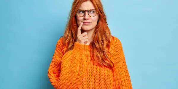 portrait-attractive-redhead-young-woman-keeps-index-finger-near-corner-lips-overthinks-something-makes-decision-wears-knitted-sweater